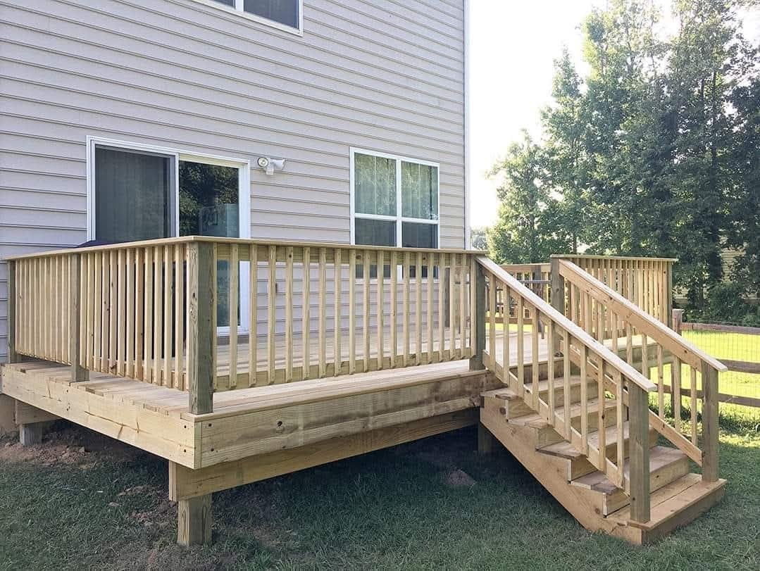 Wooden deck with railings and stairs attached to a gray residential house with green lawn and trees